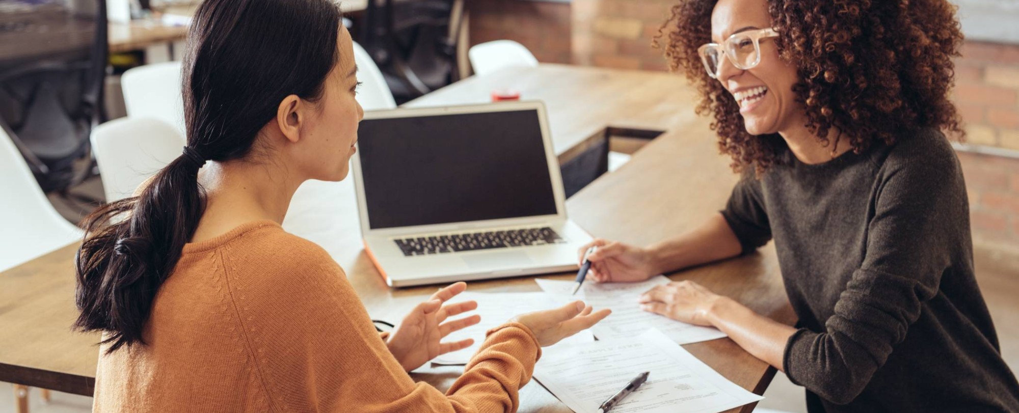 Twee vrouwen in gesprek in een kantoorsetting. Ze lachen en hebben een laptop en papieren.