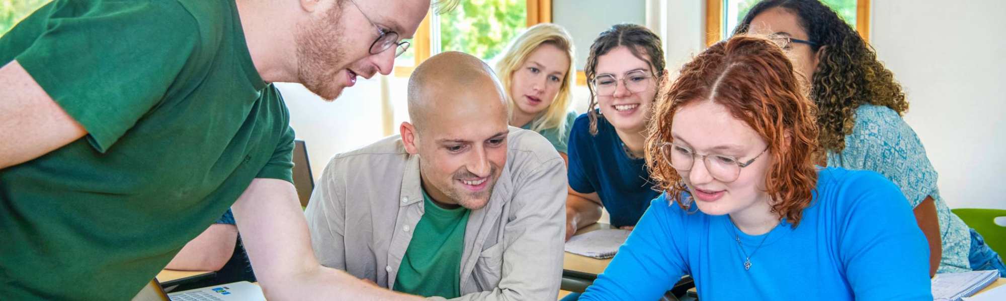 Leraar Wiskunde wijst iets aan op de laptop en klas kijkt van achter nieuwsgierig mee
