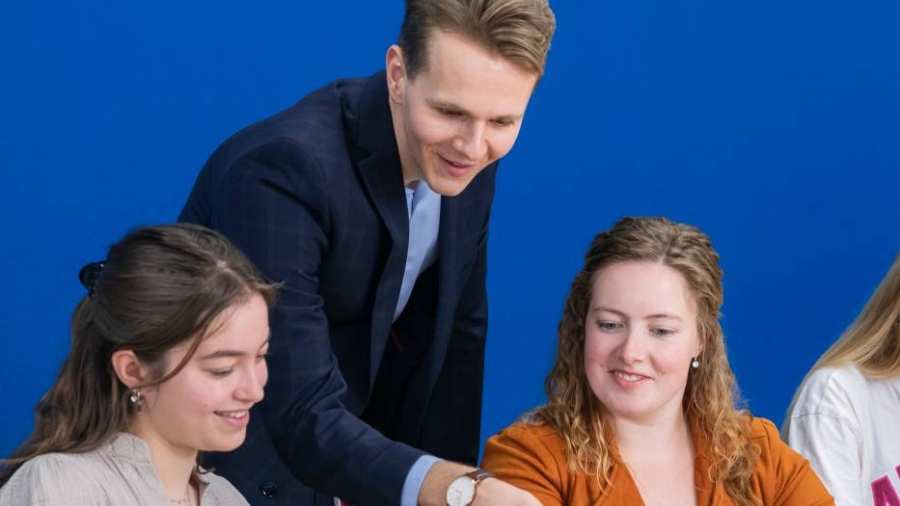 Docent in donkerblauw pak kijkt mee op de laptop bij twee van zijn studenten.
