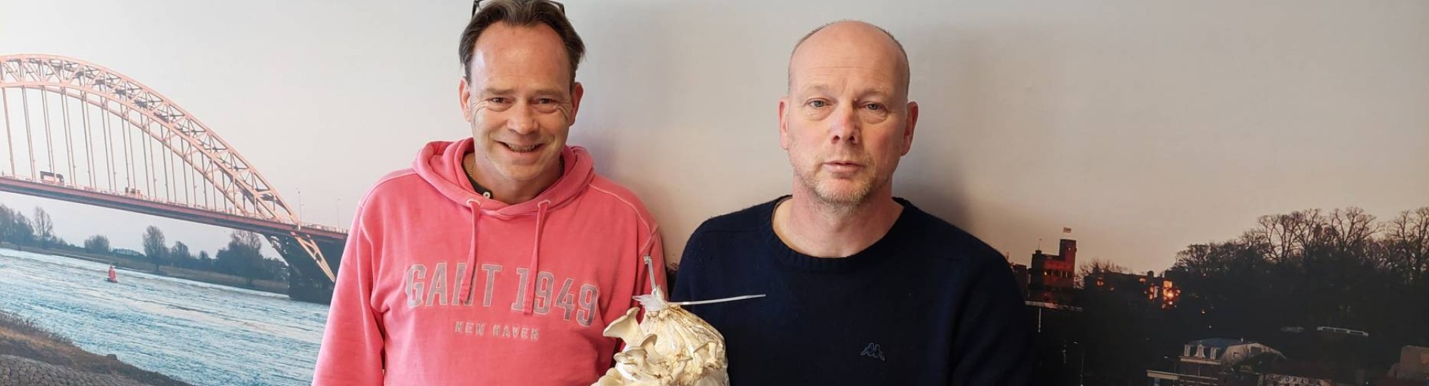 Mark Heussen en René Klep poserend voor een fotomuur met Nijmegen aan de Waal erop. Rene met oesterzwammen groeiend op een voedingsbodem van onder andere koffiedik.