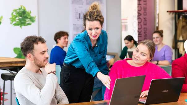 Docent helpt studenten door iets aan te wijzen op een laptop die voor de student op tafel staat. 