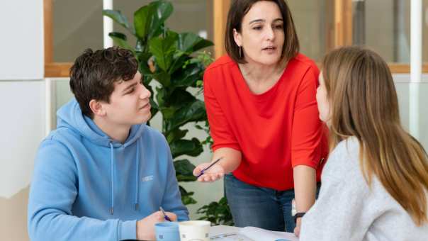 Docent geeft uitleg aan twee studenten die aan een tafeltje zitten.