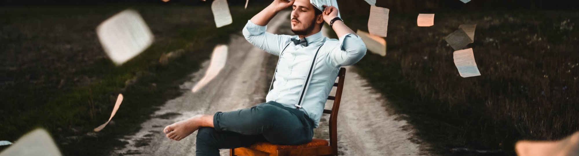 man met wit overhemd, vlinderdas en bretels zit op een stoel, op een zandweg. Hi houdt een boek boven zijn hoofd en losse pagina's vliegen in het rond