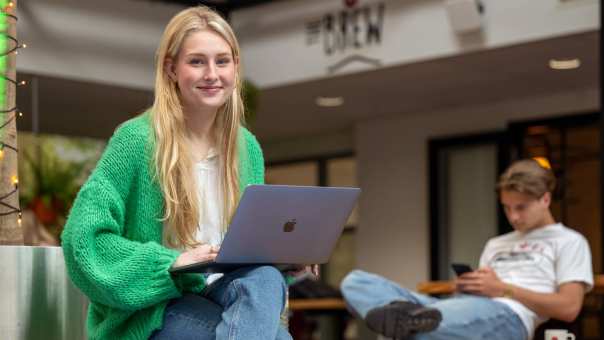 Studente lacht in de camera, heeft laptop op schoot en is aan het werk bij The Brew. 