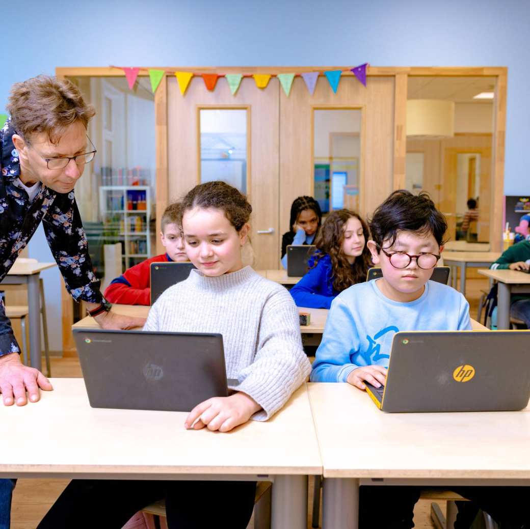 Meester legt iets uit aan een leerling en wijst naar de laptop