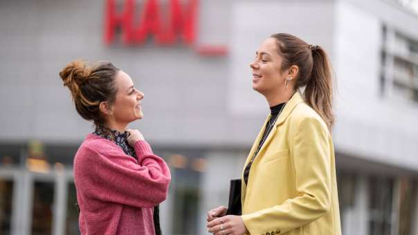 2 studenten staan buiten lachend met het HAN logo op de achtergrond. 