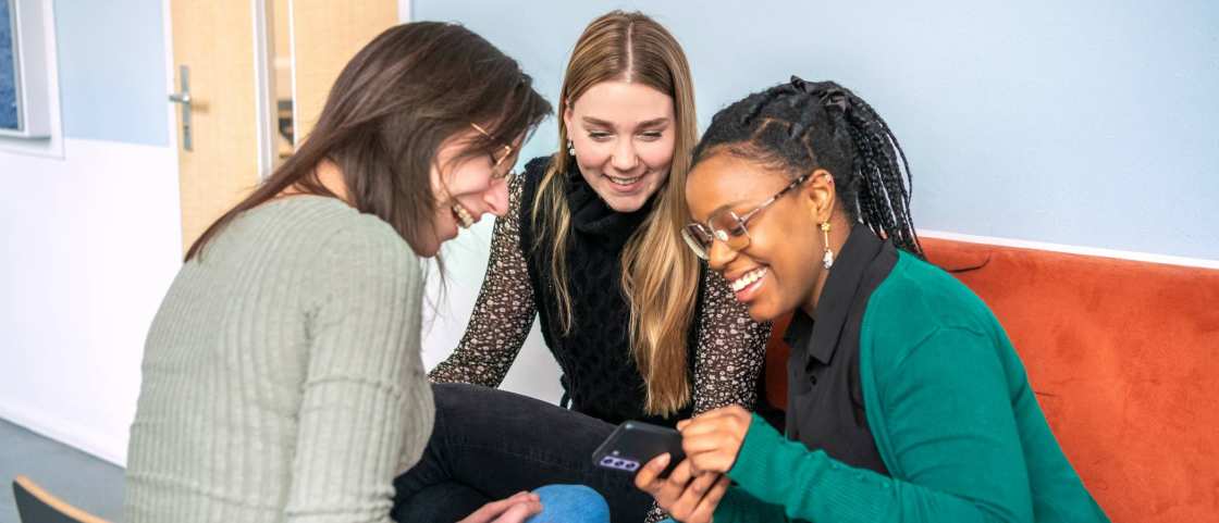 3 studenten zitten samen op een bank en kijken lachend naar een telefoon.