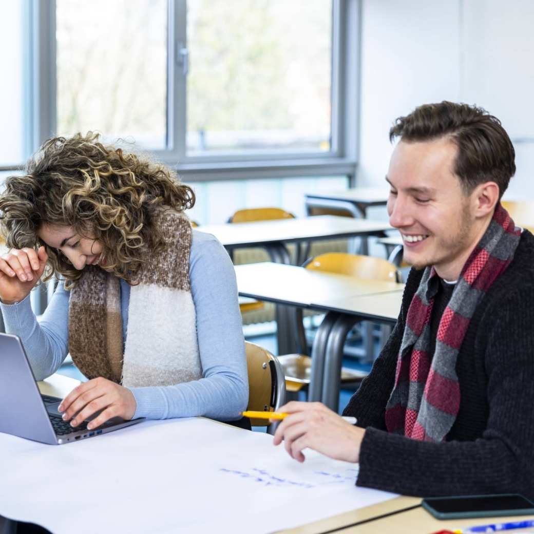 Vrouw met donkerblond krullend haar en man met donkerblond haar en zwarte trui met rode sjaal, werkend lachend aan een project met een groot wit vel papier voor zich