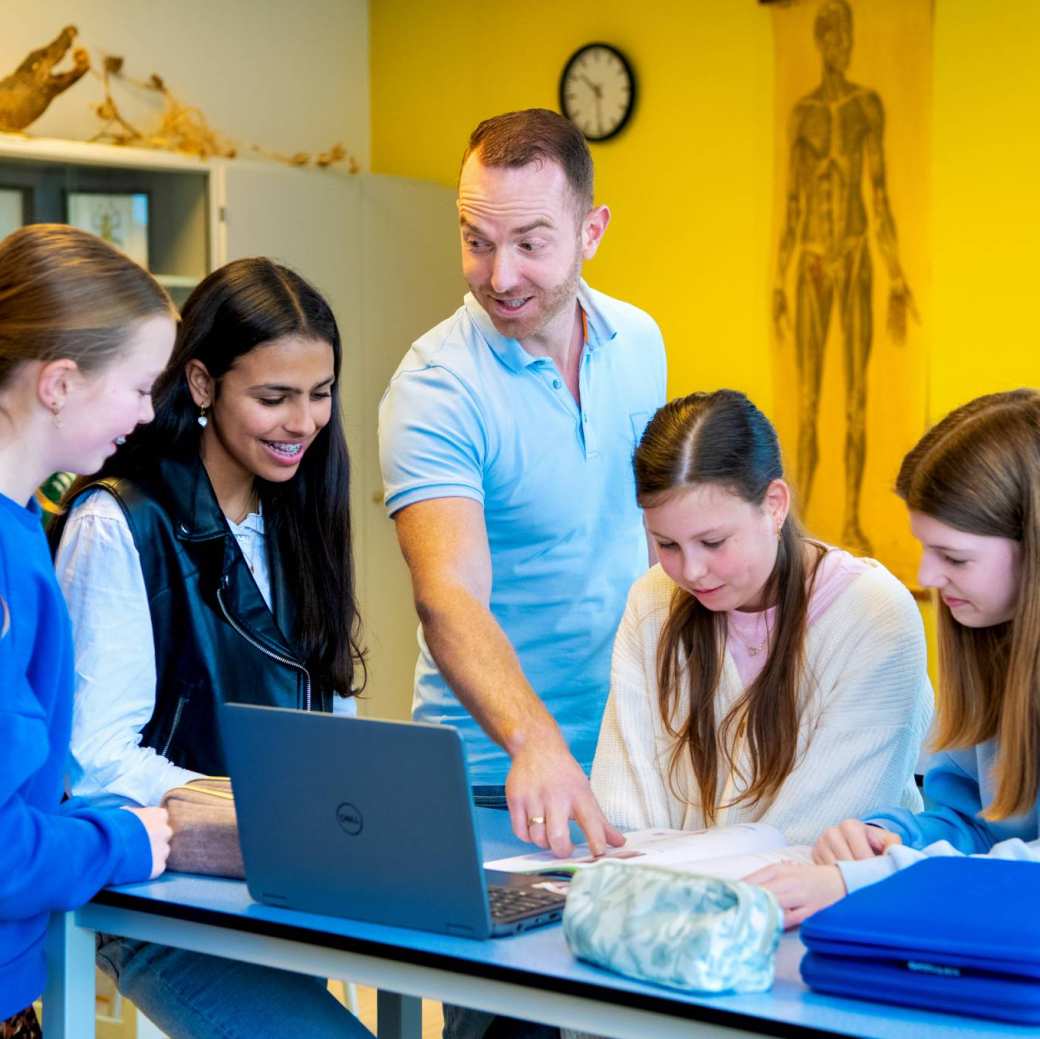 Docent geeft aan twee studenten uitleg en wijst daarbij naar de pagina in het boek dat de studenten in de hand hebben.