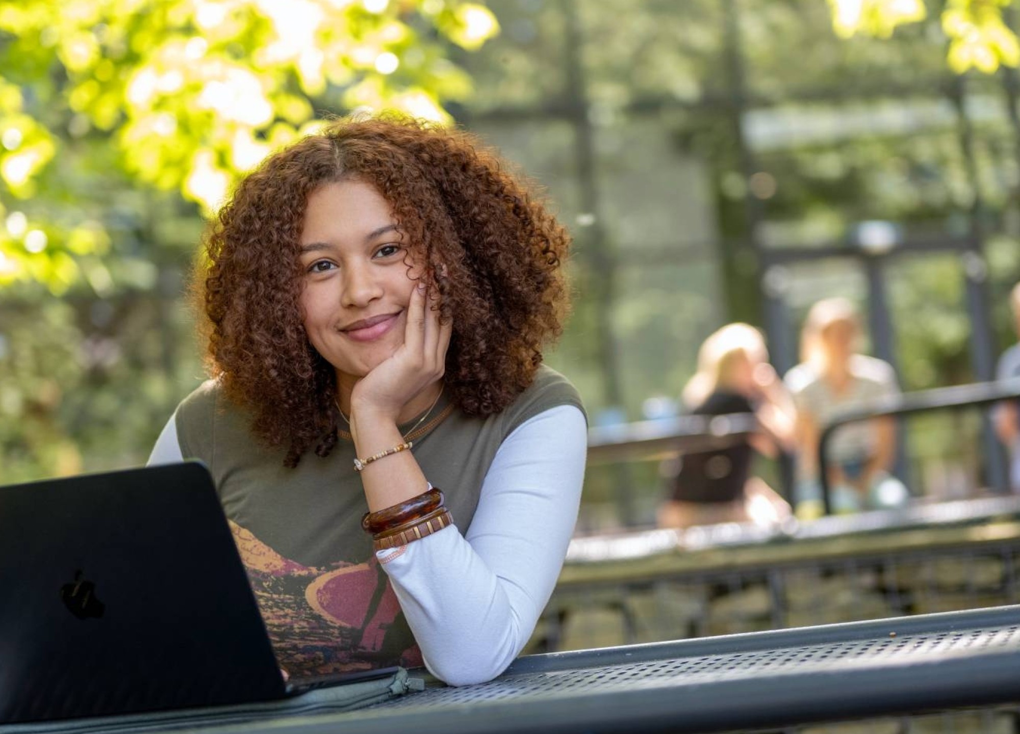 Studente kijkt in de camera, hand onder haar hoofd, werkt op laptop, buiten op bankje bij K33. 