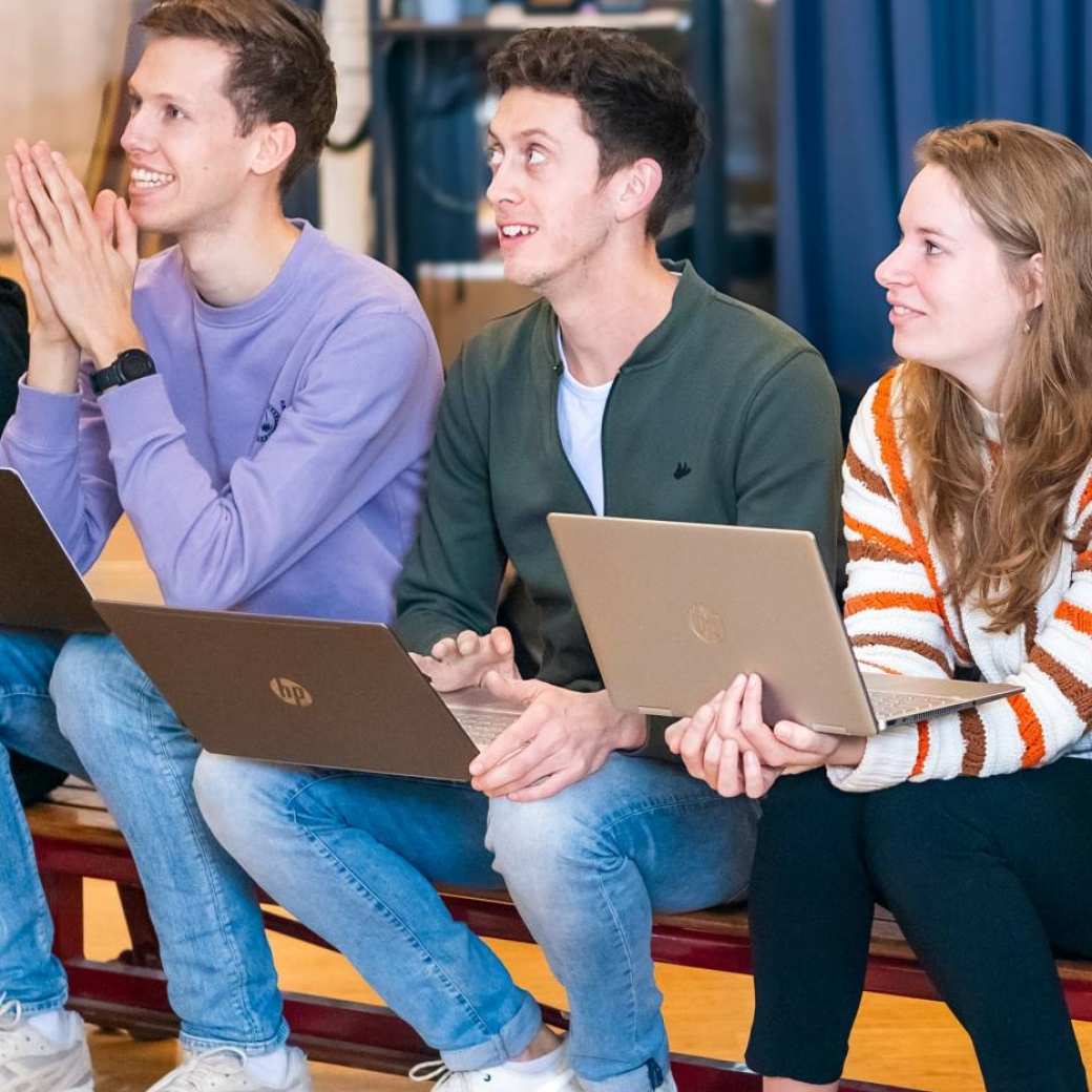 Studenten zitten op een gymbank, kijken erg hoopvol. 