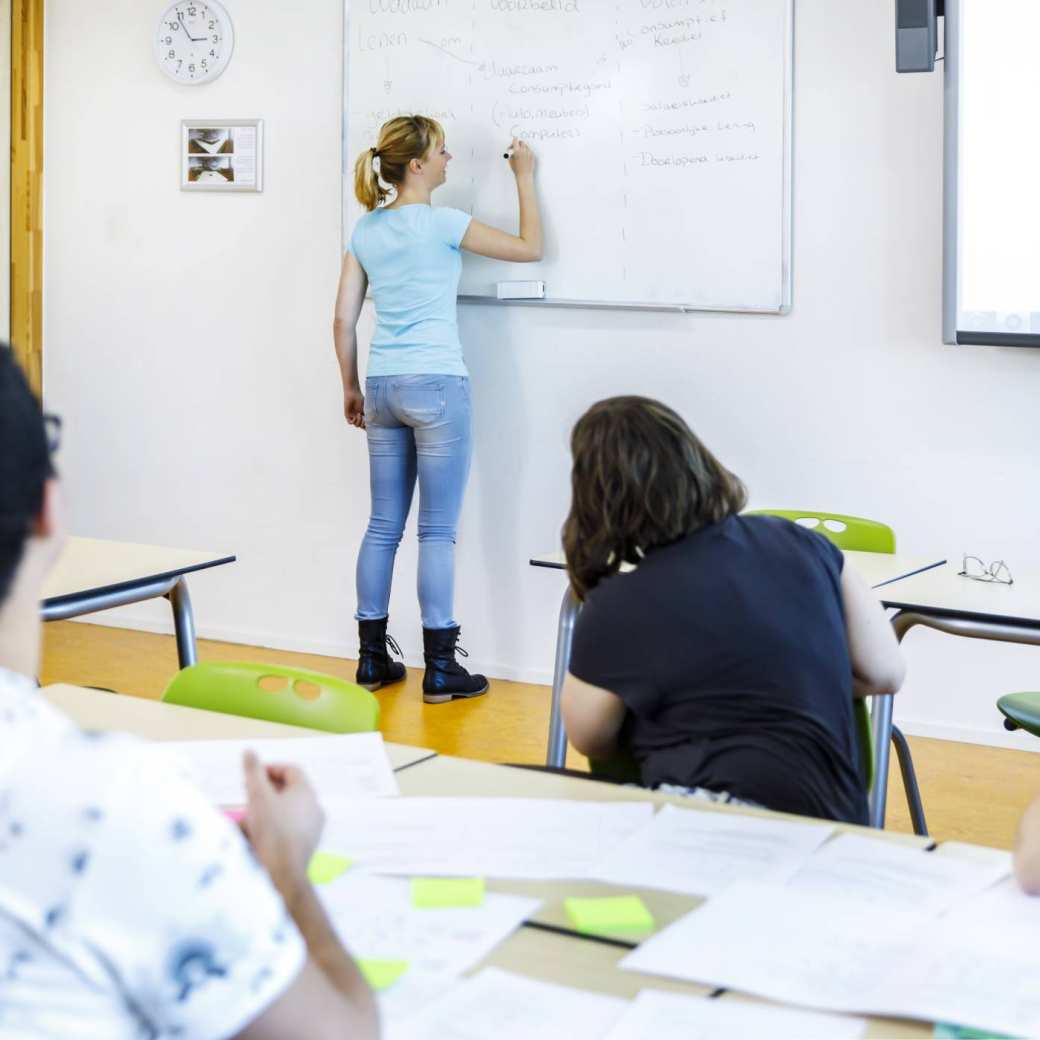 Foto van achterin de klas gemaakt van een student die met marker op het whiteboard schrijft. 