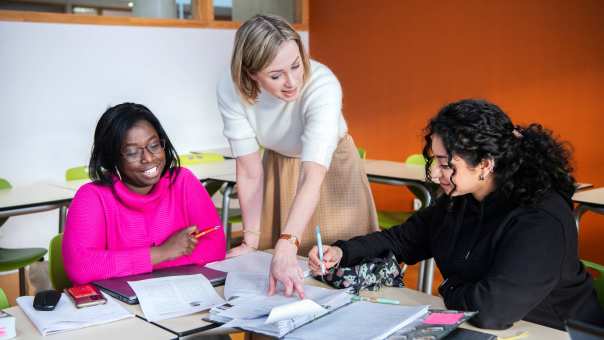 docent wijst in een boek terwijl ze bij twee studentes staat