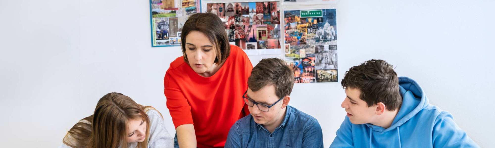 Drie studenten zitten op een rij en de docent staat achter hen. Ze wijst in het boek en de studenten kijken mee.