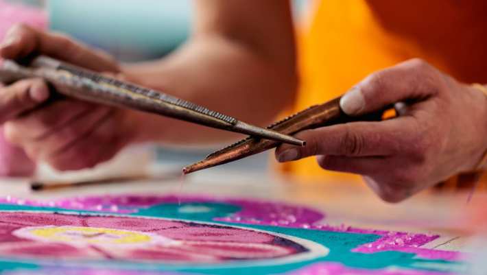 Persoon in oranje gewaad maakt met metalen gereedschap een kleurrijke zand mandala