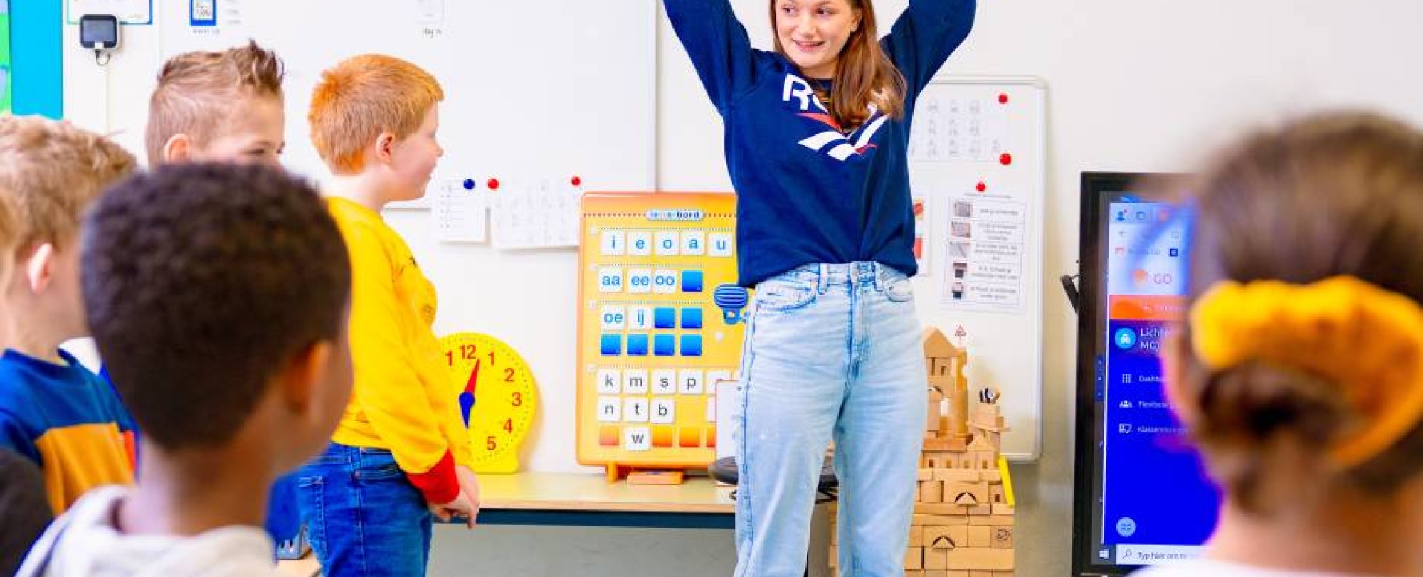 Juf staat voor de klas en steekt haar handen in de lucht