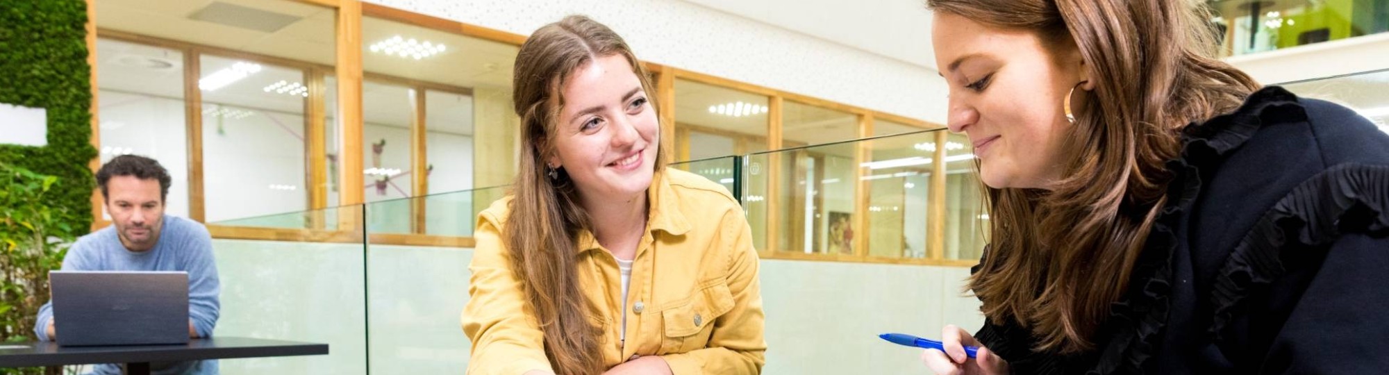 ALPO-student zit samen met haar docent te sparren aan een tafeltje in de gang van het I/O gebouw. 