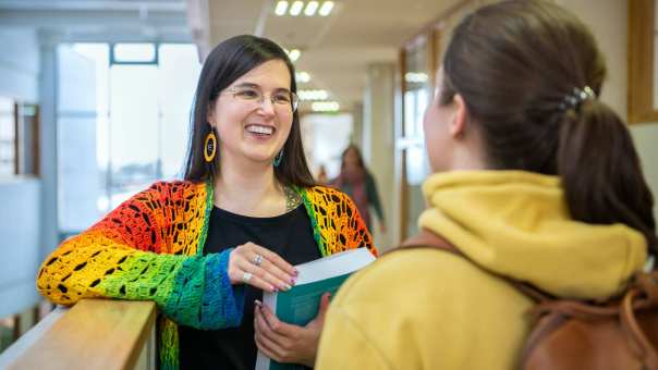 2 masterstudenten staan lachend in de gang met elkaar te praten met een studieboek in de hand