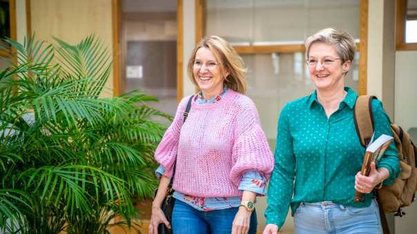 Twee studenten lopen naast elkaar en lachen, terwijl ze door de gang lopen.