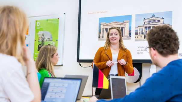 Student leraar Duits in oranje/bruin jasje geeft een lesje aan haar medestudenten. Ze staat voor een PowerPoint.
