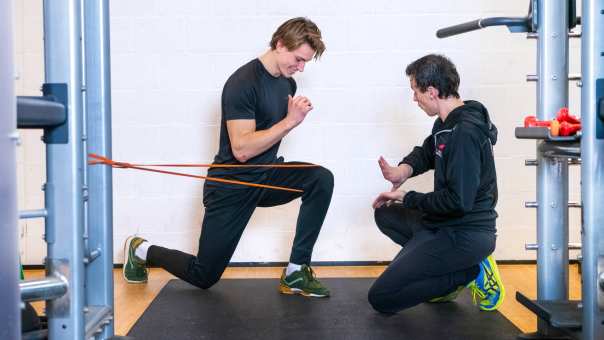 Student en docent oefenen in gymzaal, jongen zit op 1 knie en stretcht zijn been. 