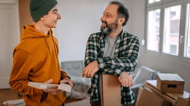 Vader met grijzend baardje leunt op een kartonnen doos. Hij praat met zijn zoon met zwarte muts die een lattenbodem van een bed vasthoudt in de studentenkamer waar hij gaat wonen.