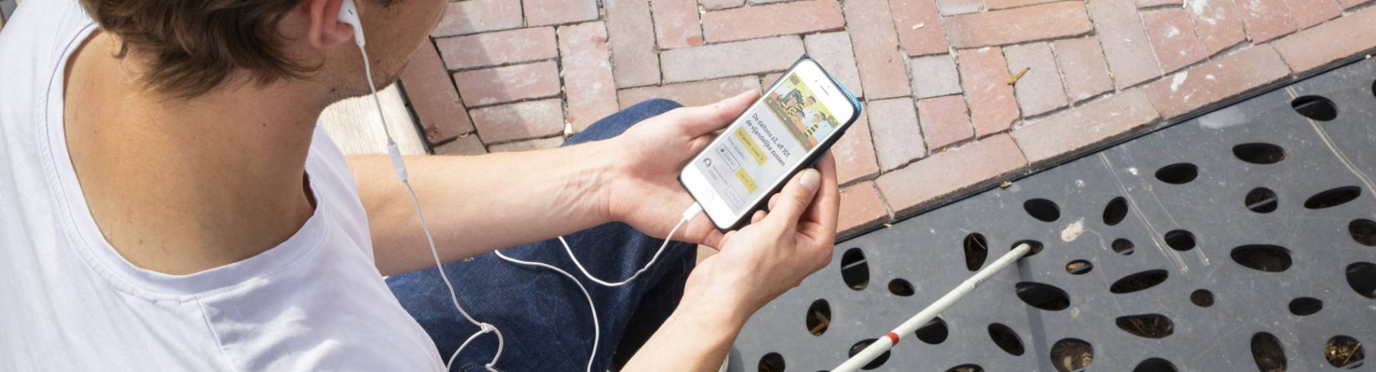 Jongen zit op een bankje en is met zijn mobiele telefoon bezig. Voor hem ligt zijn taststok.