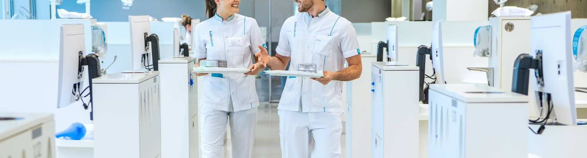 2 studenten kijken elkaar aan en lopen door een ruimte met veel behandelstoelen.