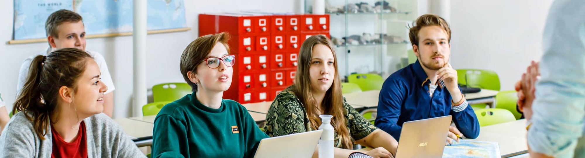 5 studenten zitten aan tafel en kijken en luisteren naar de uitleg van de docent voor hun