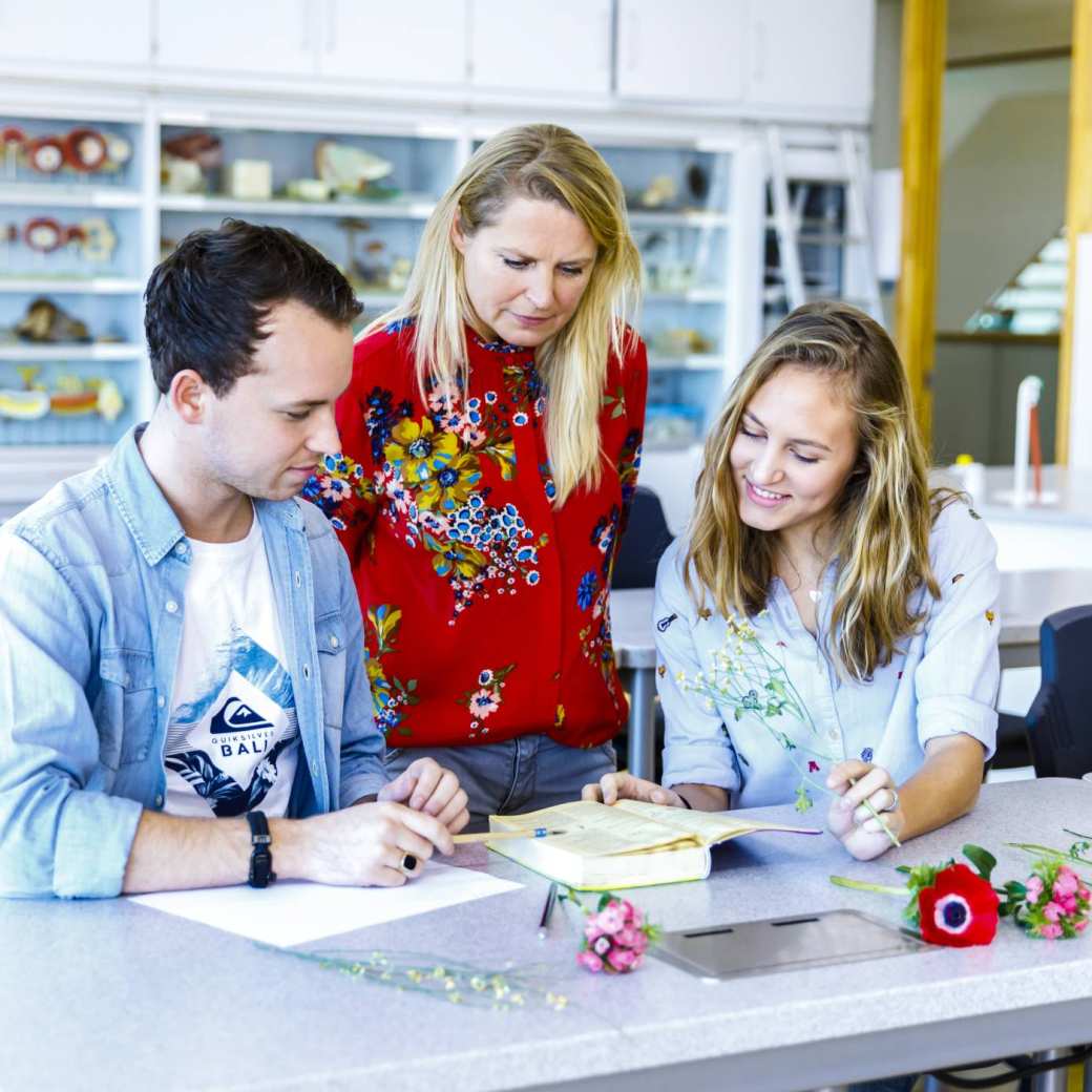 Twee studenten maken een opdracht en vragen daar iets over aan de docent die kritisch nadenkt. 