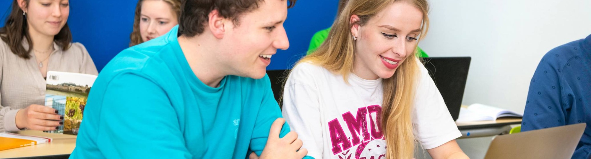 Docent geeft aan twee studenten uitleg en wijst daarbij naar de pagina in het boek, dat de studenten in de hand hebben.