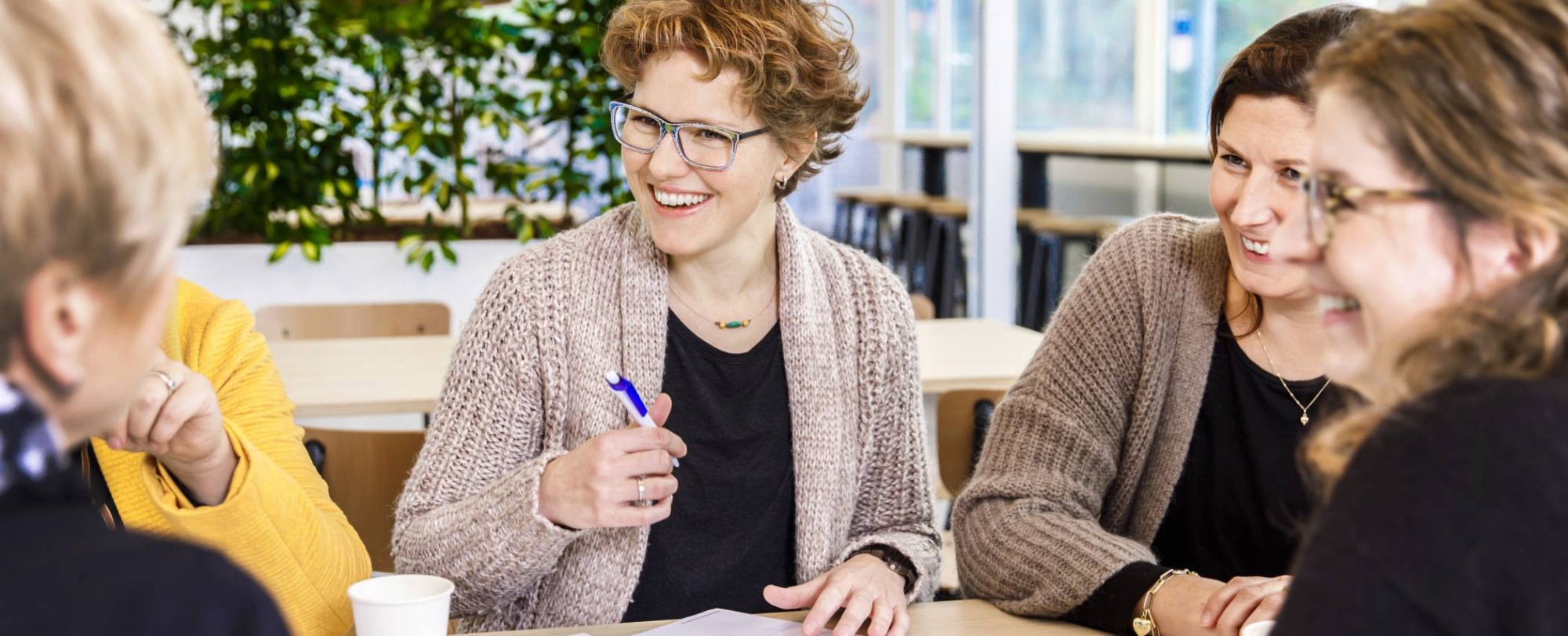 Vijf vrouwen zijn met elkaar aan het overleggen aan een tafel. Een van de vrouwen maakt notities.