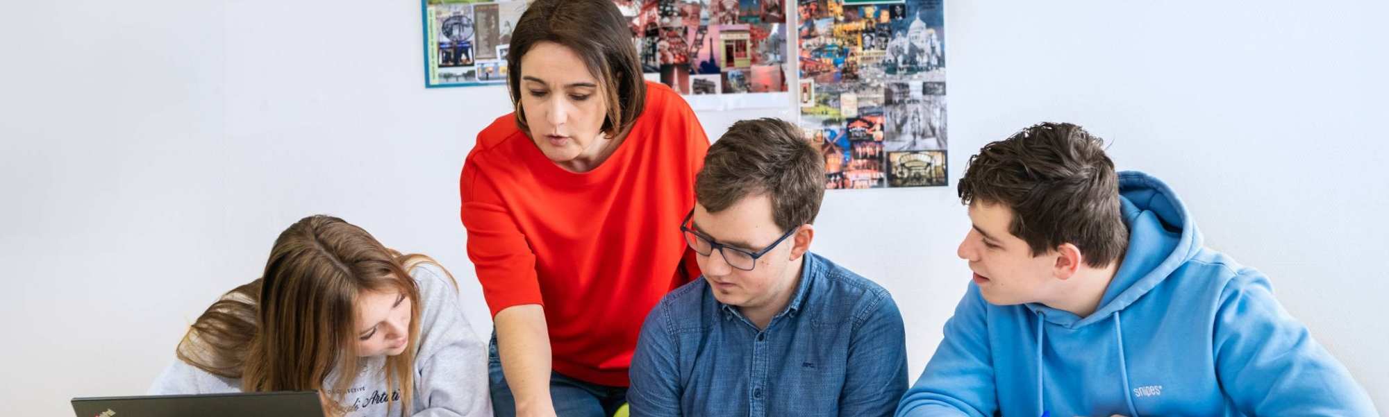 Drie studenten zitten op een rij en de docent staat achter hen. Ze wijst in het boek en de studenten kijken mee.