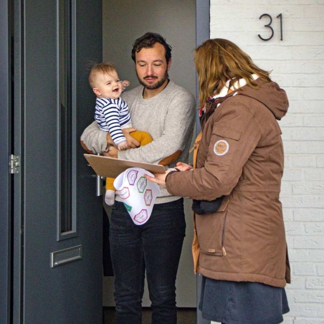 Een man met kind op de arm in de deurpost, in gesprek met een dame die veldonderzoek doet.