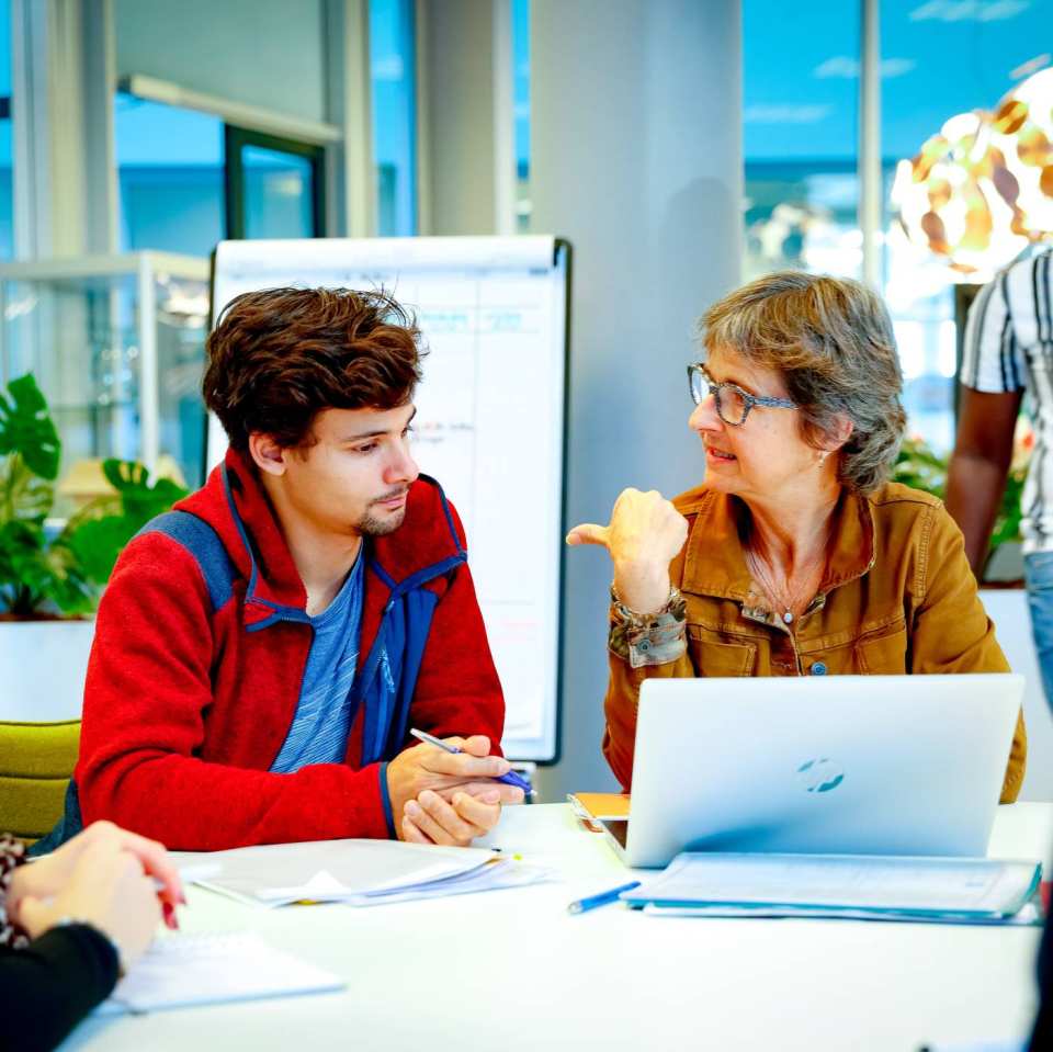 Docent zit met een laptop en drie studenten om tafel en wijst de student naast haar aan. 