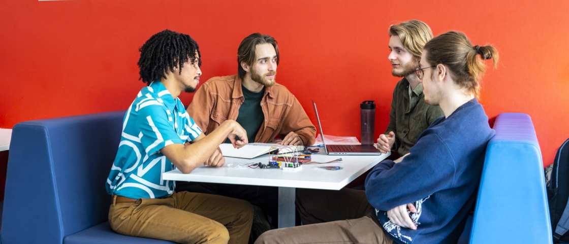 Studenten en docent in overleg met elkaar, bij blauwe banken en rode muur. 