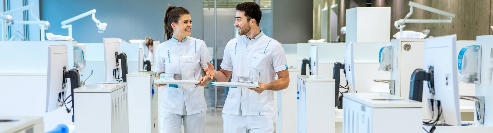 2 studenten kijken elkaar aan en lopen door een ruimte met veel behandelstoelen.