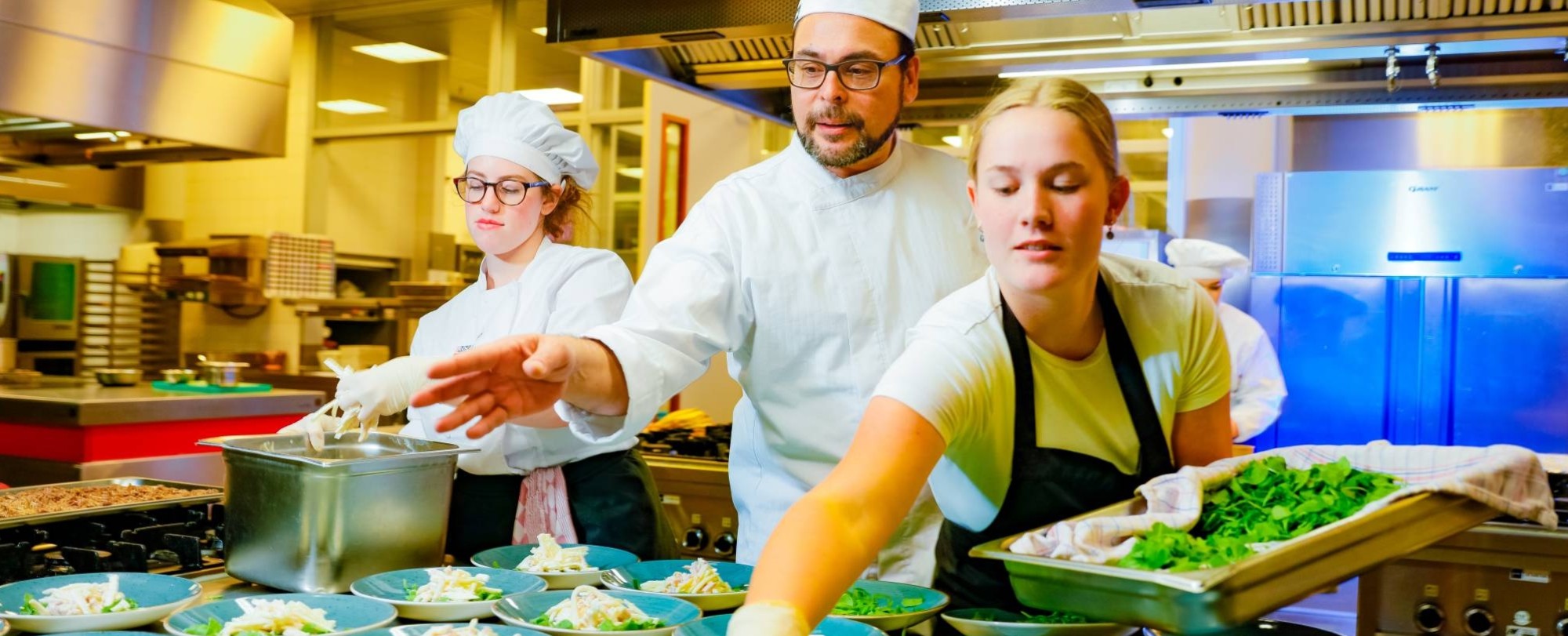 Student legt sla op een bord terwijl de chefkok naast haar instructies geeft.