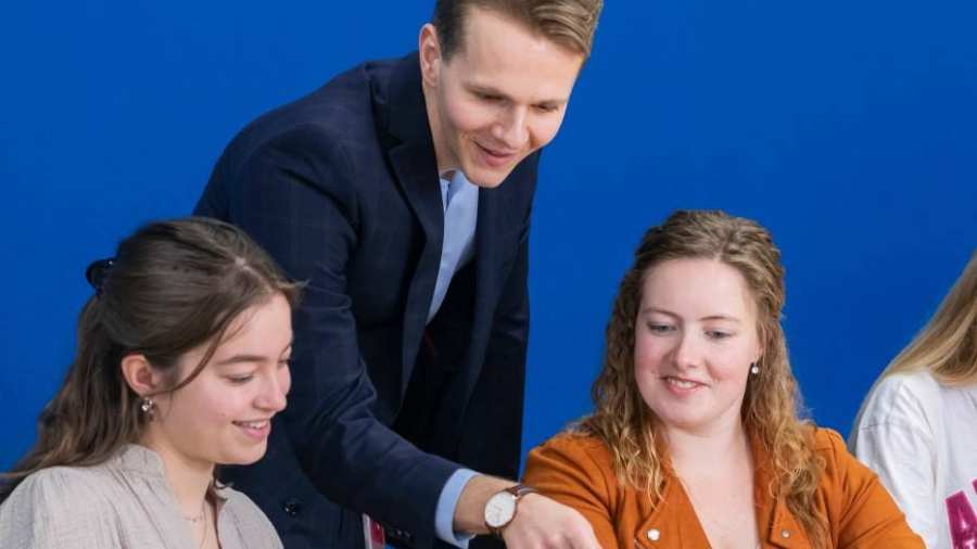 Docent in donkerblauw pak kijkt mee op de laptop bij twee van zijn studenten.
