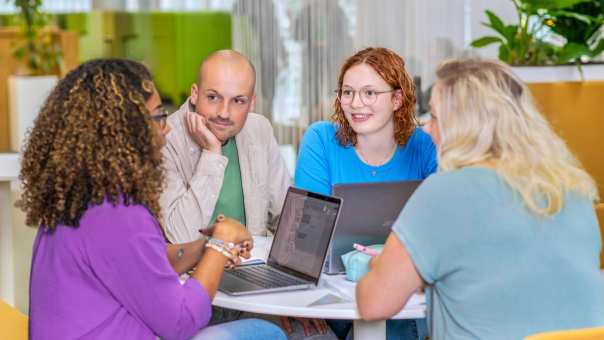 Vier studenten Leraar Wiskunde overleggen aan een ronde tafel en luisteren aandachtig naar elkaar. Op de tafel liggen etuis, schriften en laptops.
