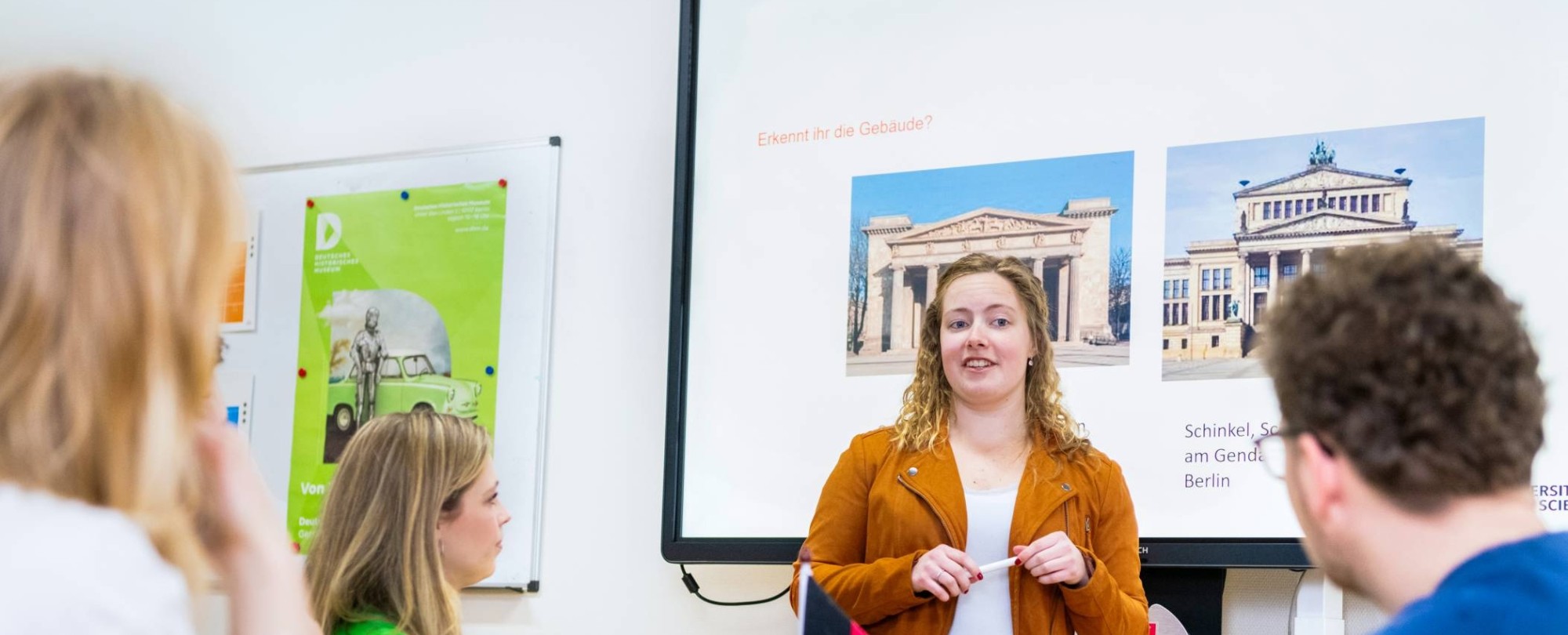 Docent geeft aan twee studenten uitleg en wijst daarbij naar de pagina in het boek dat de studenten in de hand hebben.