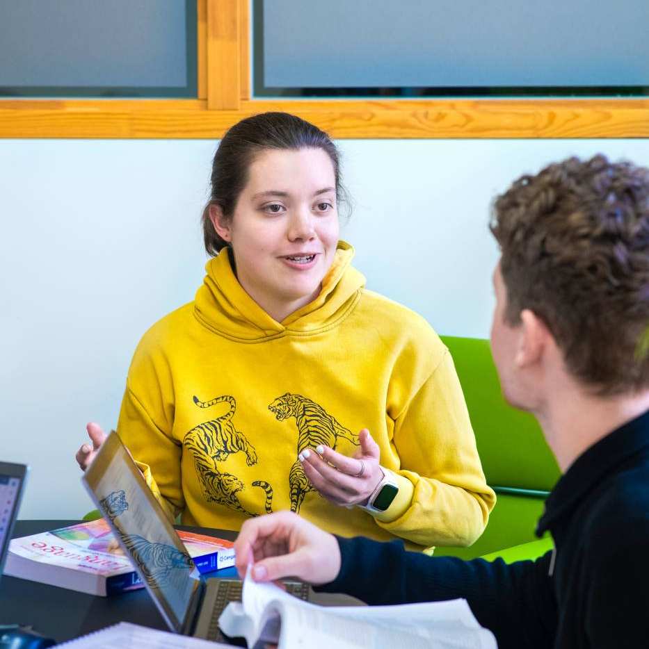 Foto Master Academie Educatie. 2 masterstudenten zijn met elkaar in gesprek aan een tafel met laptops en boeken. 