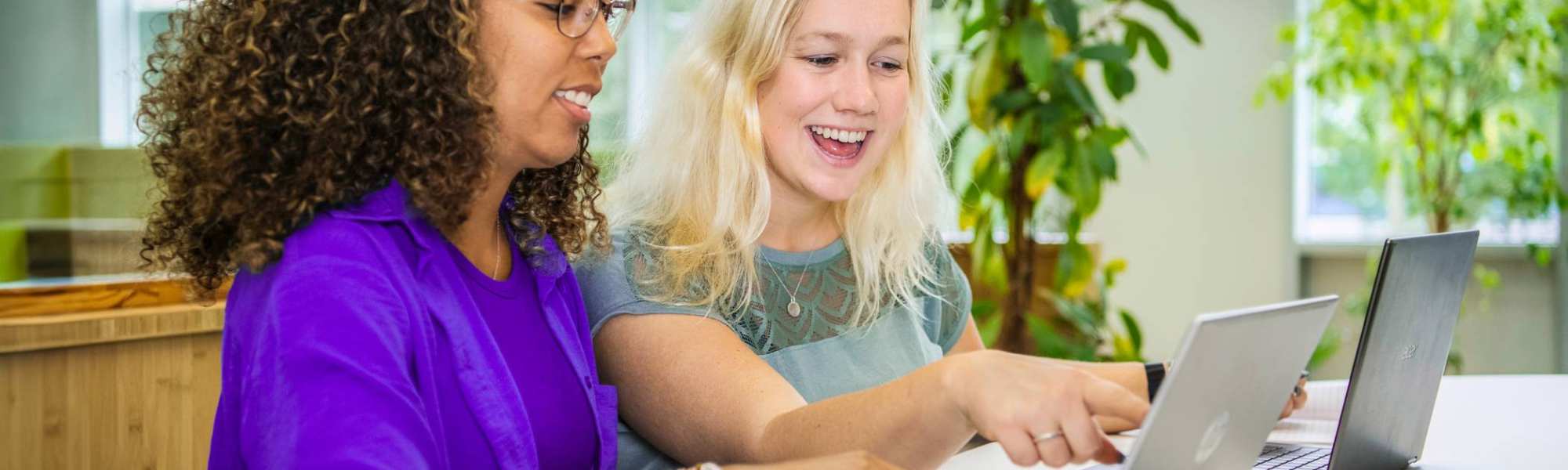 Twee studenten Leraar Wiskunde zitten naast elkaar met een laptop. De ene wijst lachend op het scherm iets aan en de ander kijkt lachend mee.
