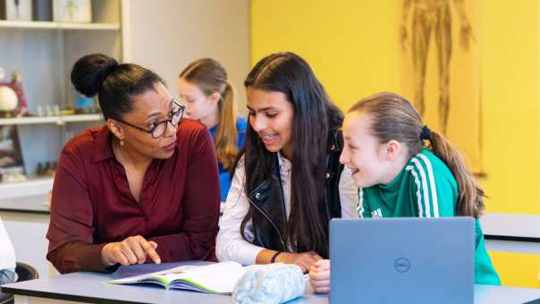 Docenten Claudine Laclé geeft uitleg aan 2 leerlingen en wijst in het schrift voor ze op tafel op het Citadel College. 