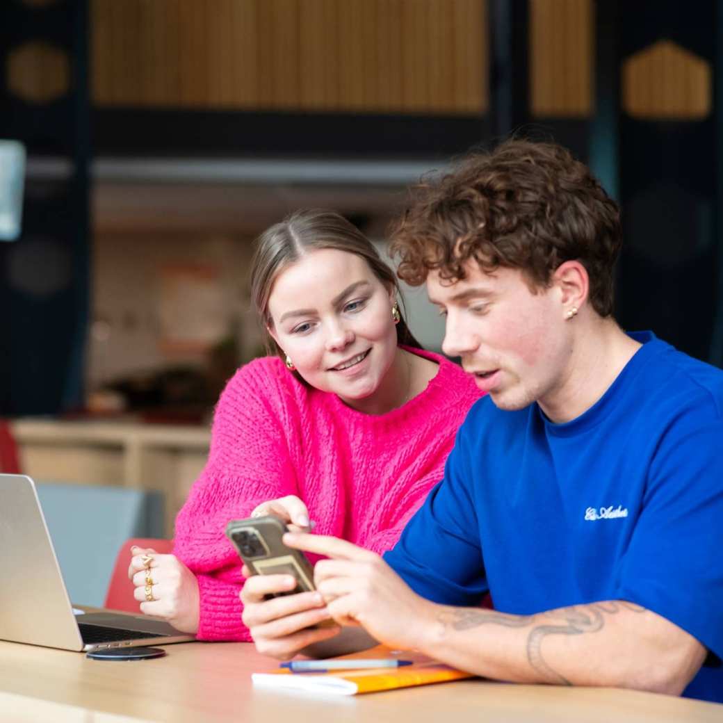 Studenten zitten samen aan tafel en kijken op 1 telefoon.