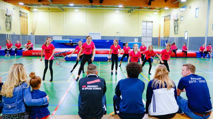 Ouders en bezoekers zitten op bankje in gymzaal en kijken naar sport en dans