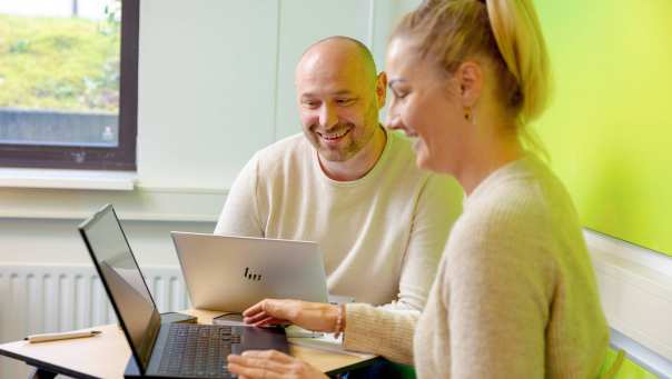 Vrouw en man zitten achter laptop, allebei aan het lachen. Groene muur op achtergrond.