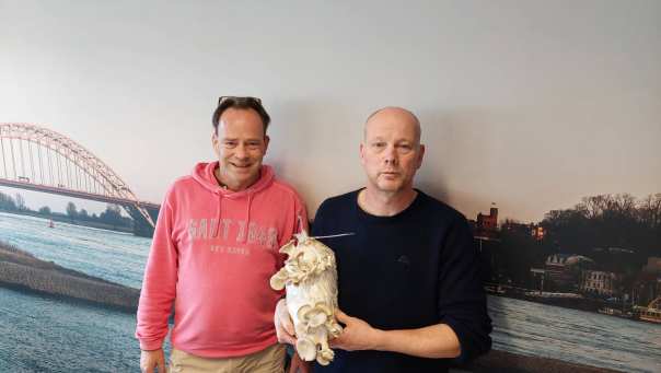 Procescoördinator Mark Heussen en Ruud Klep van de Versfabriek poseren voor een afbeelding van Nijmegen aan de Waal met een zak waarop oesterzwammen groeien op mengsel met koffiedrab.