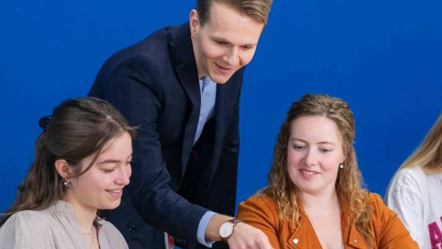 Docent in donkerblauw pak kijkt mee op de laptop bij twee van zijn studenten.
