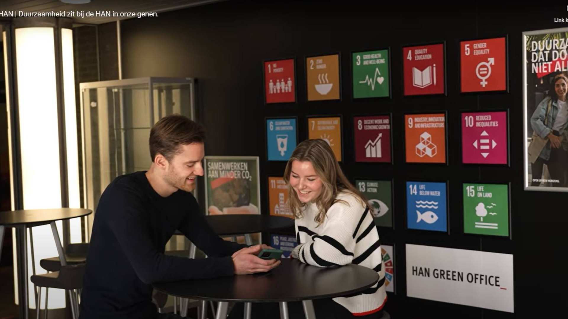 Still van de wand van de Green Office met alle SDG's en twee studenten van de GO ervoor.